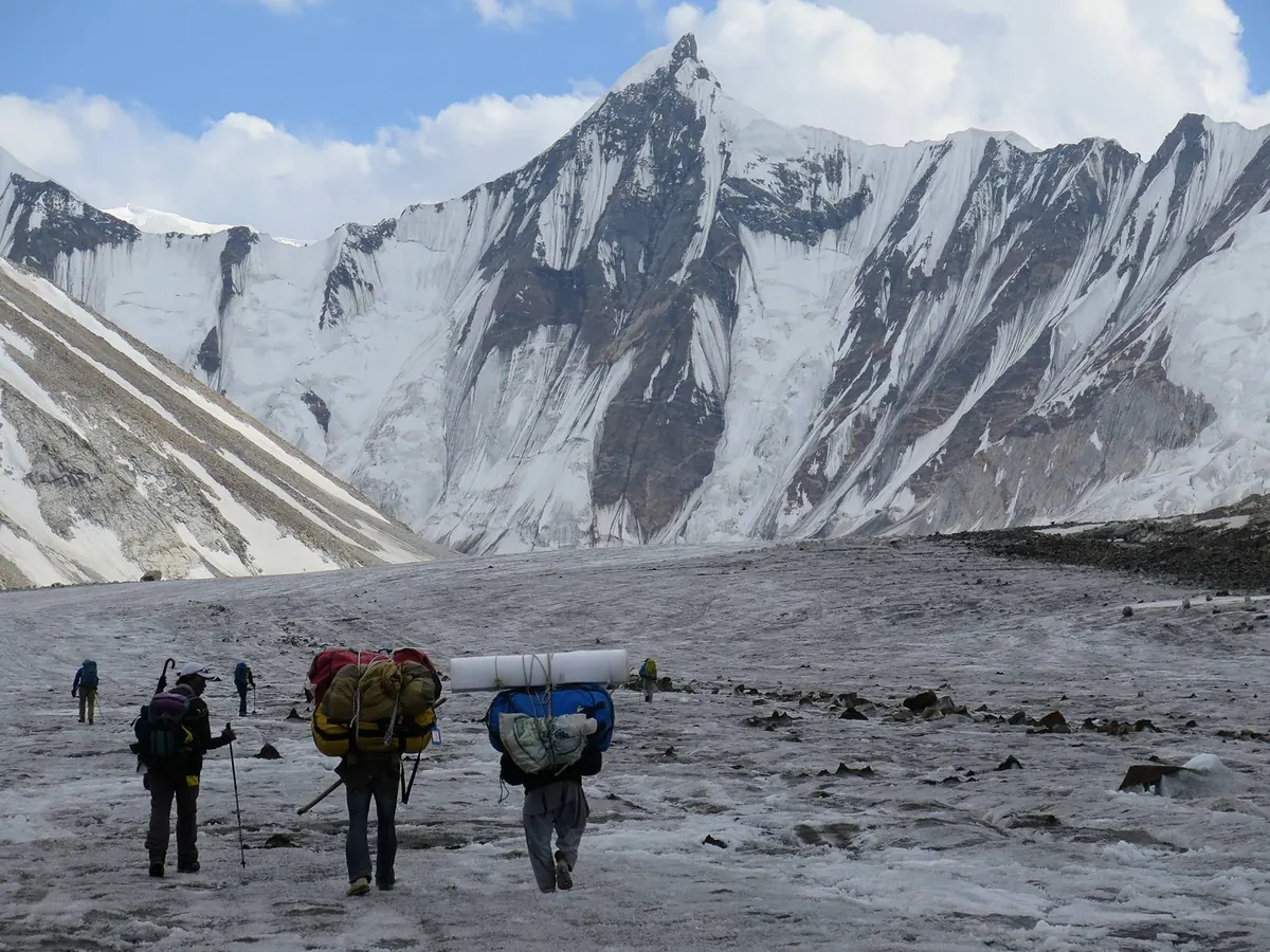 Trekker und Träger auf dem Gondogoro-Gletscher auf dem Weg zum Ali Camp unter einem verschneiten Granitgipfel, Hushe-Seite des Gondogoro La, Pakistan