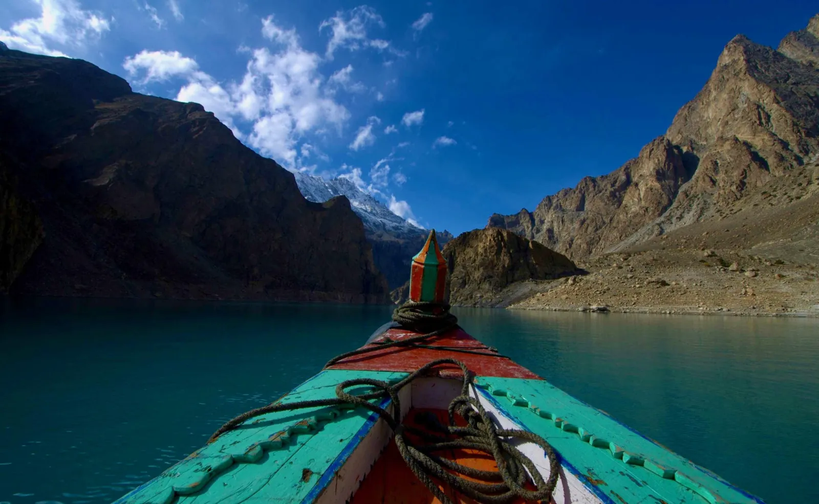 First-person view from the bow of a colourful wooden boat gliding across the turquoise water of Attabad Lake toward snow-capped Karakoram peaks, Upper Hunza, Pakistan