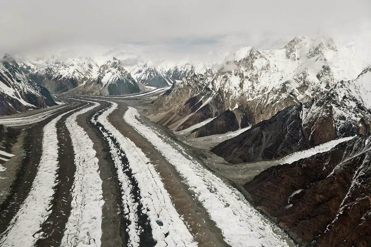 Dunkle Moränenstreifen auf dem Baltoro-Gletscher zwischen verschneiten Karakorum-Gipfeln, Gilgit-Baltistan, Pakistan