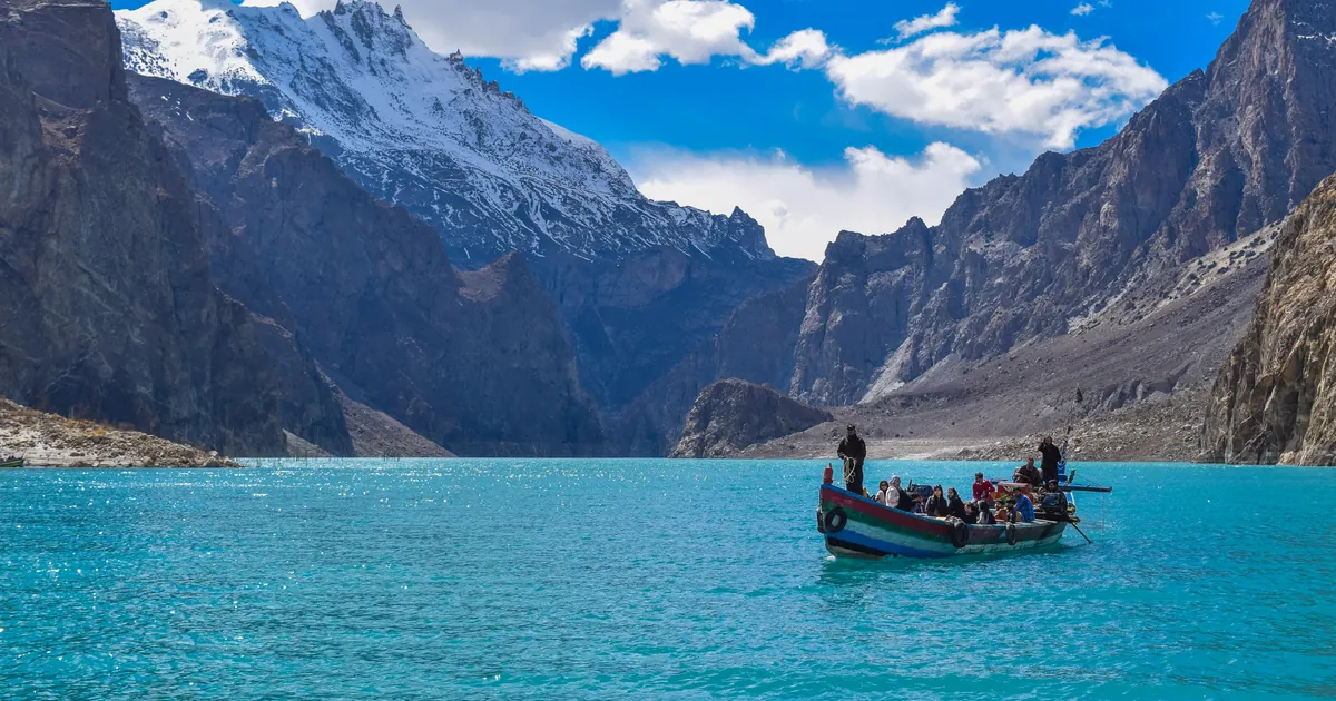 A wooden boat on the turquoise water of Attabad Lake in Hunza, below a snow-capped peak and steep grey cliffs