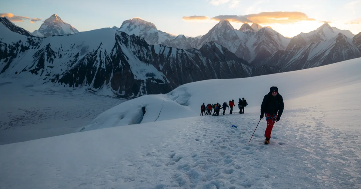 A roped line of trekkers ascends a dawn-lit snowfield beneath K2 and the Karakoram giants on the Gondogoro La Trek in Pakistan