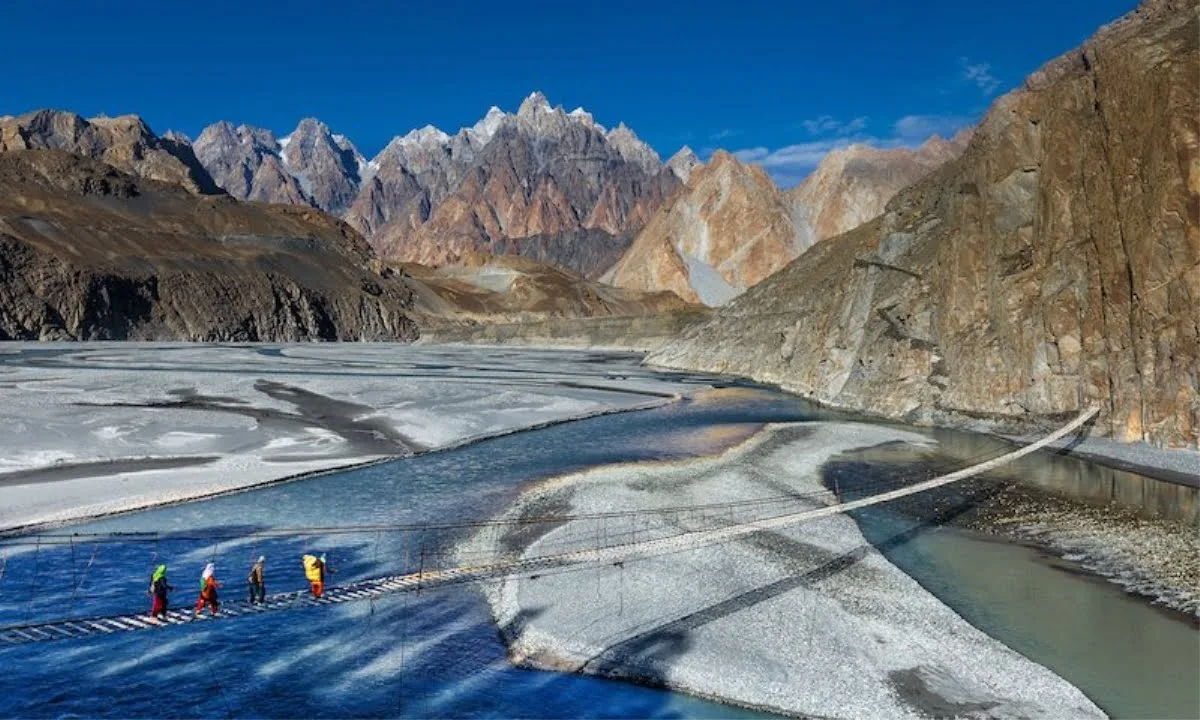 Escursionisti che attraversano il ponte sospeso a listelli di Hussaini sopra il fiume Hunza vicino a Passu, con i frastagliati Passu Cones sullo sfondo