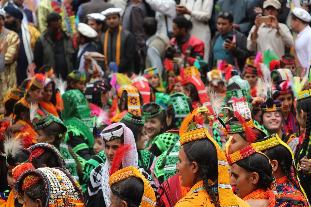 Kalash women in green, orange and cowrie-shell headdresses dancing at a festival while a crowd of visitors watches and films behind them