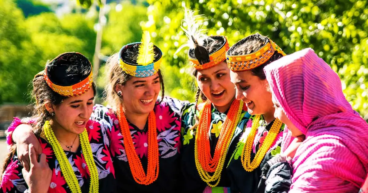 Five Kalash women in beaded feather headdresses and orange and yellow necklaces laughing together at a festival in the Kalash valleys, Chitral