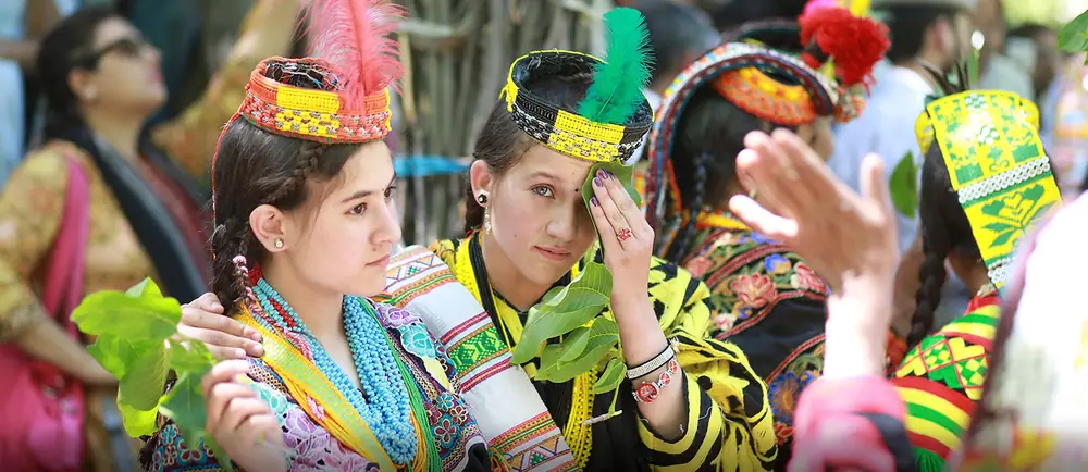 Two Kalash girls in feathered beaded headdresses holding green walnut leaves during the Chilam Joshi spring festival