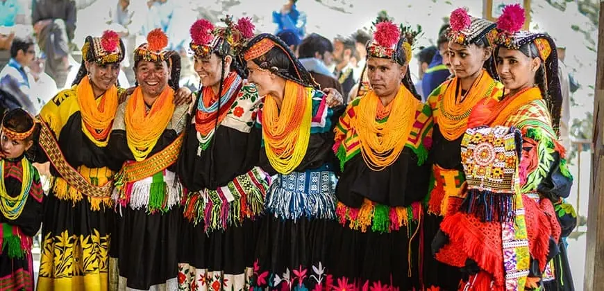 A line of Kalash women in black embroidered dresses with orange and yellow beaded necklaces and flower-topped headdresses at a festival