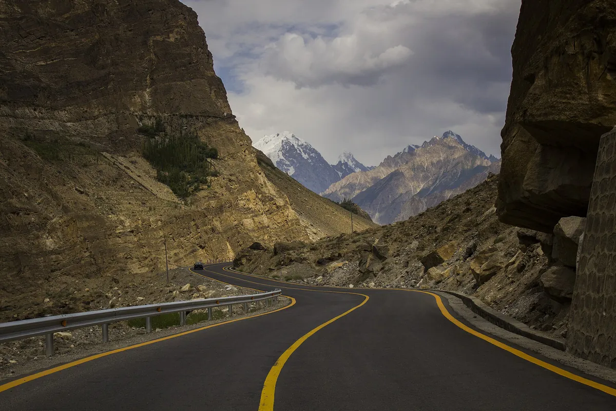 The Karakoram Highway curving through a rock cutting near Jaglot, Gilgit, with snow peaks ahead on the road to Skardu