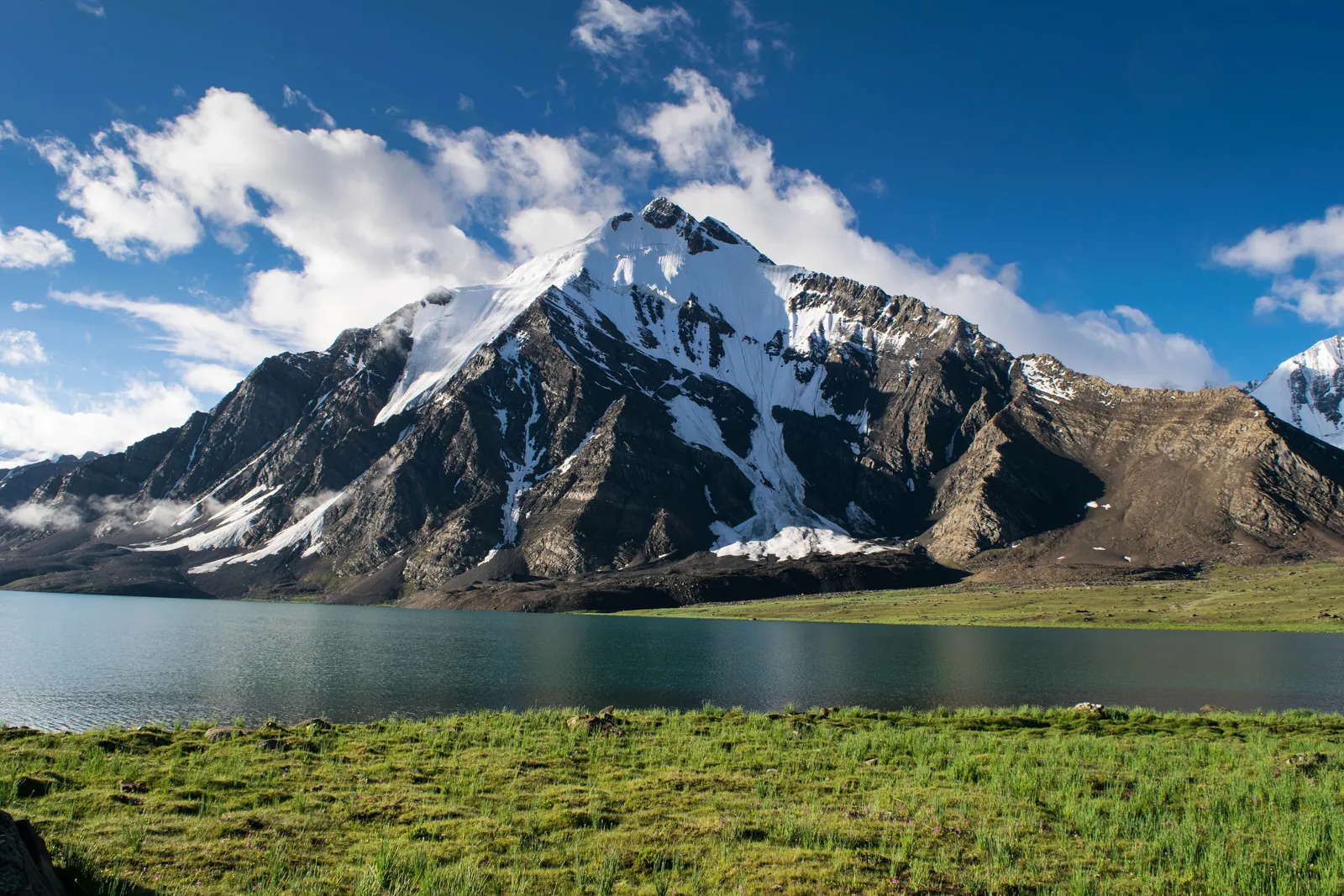 Karambar Lake below a snow-covered peak, with green grass along the shore, on the Broghil and Ishkoman border