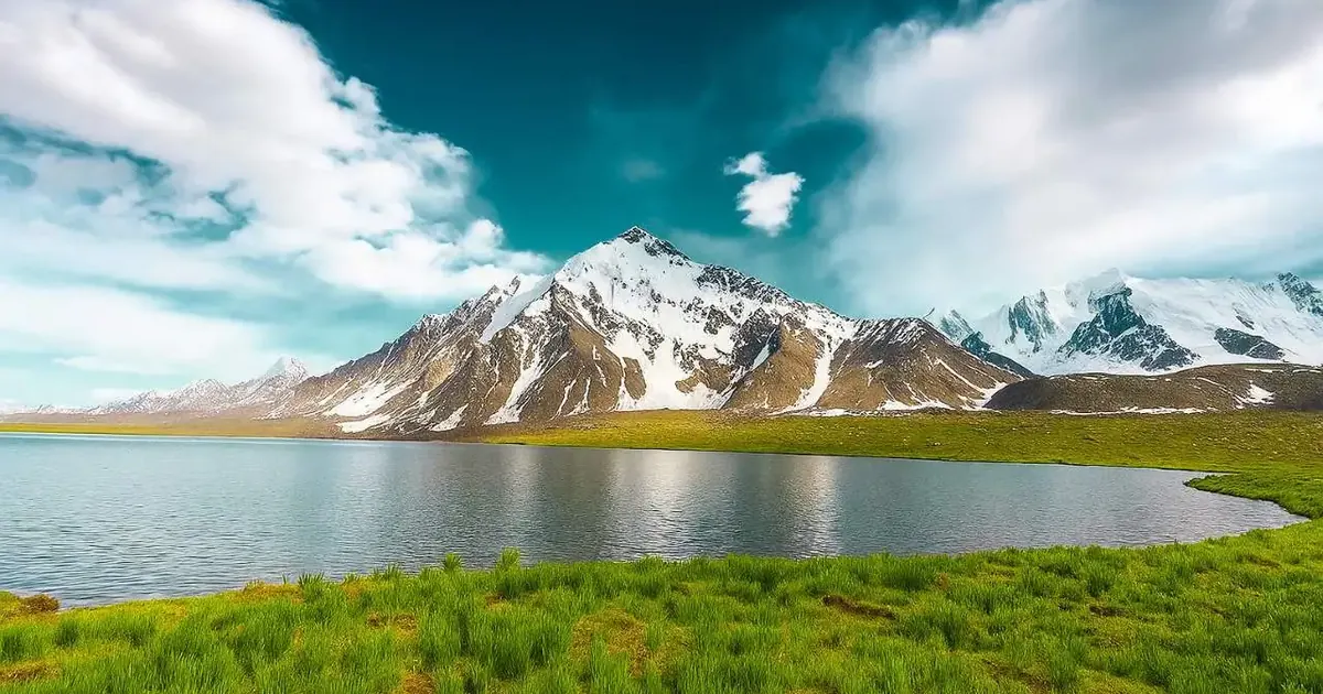 Karambar Lake at 4,272 m with snow-covered peaks reflected in still water and green summer pasture along the shoreline, Broghil, Pakistan