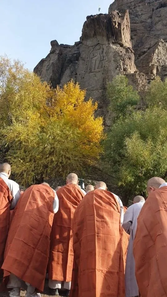 Buddhist monks in orange robes standing below the Kargah Buddha and looking up at the carving, with autumn poplars behind them