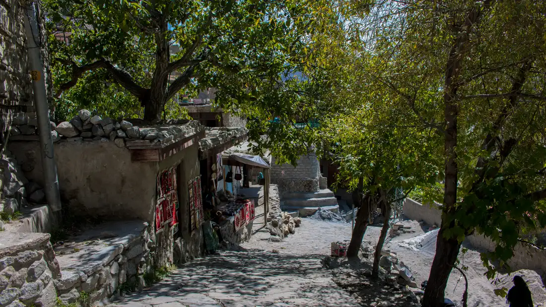 Vicolo con muretti in pietra davanti a una piccola bottega di artigianato all'ombra degli alberi a Karimabad, valle di Hunza, Pakistan