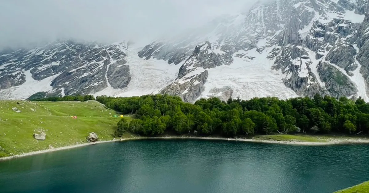 Kutwal Lake in the Haramosh Valley, with camp tents on the meadow, a treeline of willow and birch, and the ice face of Haramosh Peak behind