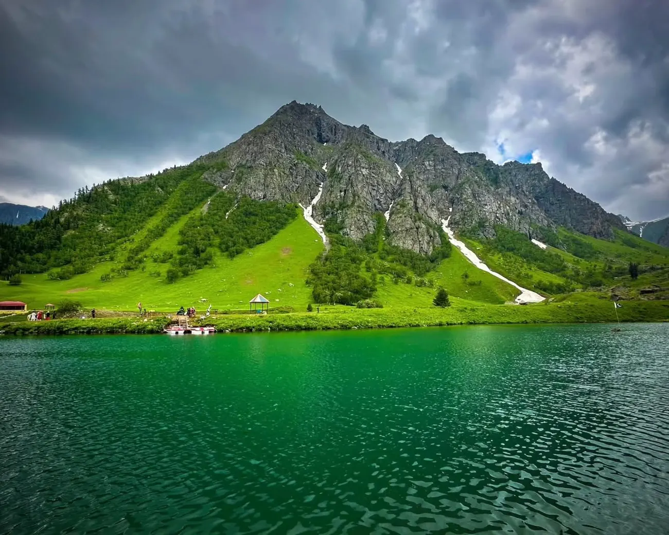 A deep green alpine lake below a steep pyramid peak in the Astore valley under a broken sky
