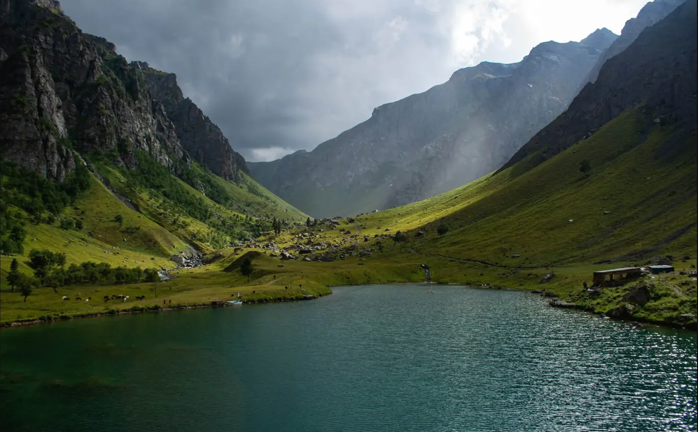 A green alpine lake below steep grassy walls near Domel in the Minimarg area of Astore, with grazing cattle and a single building on the far shore