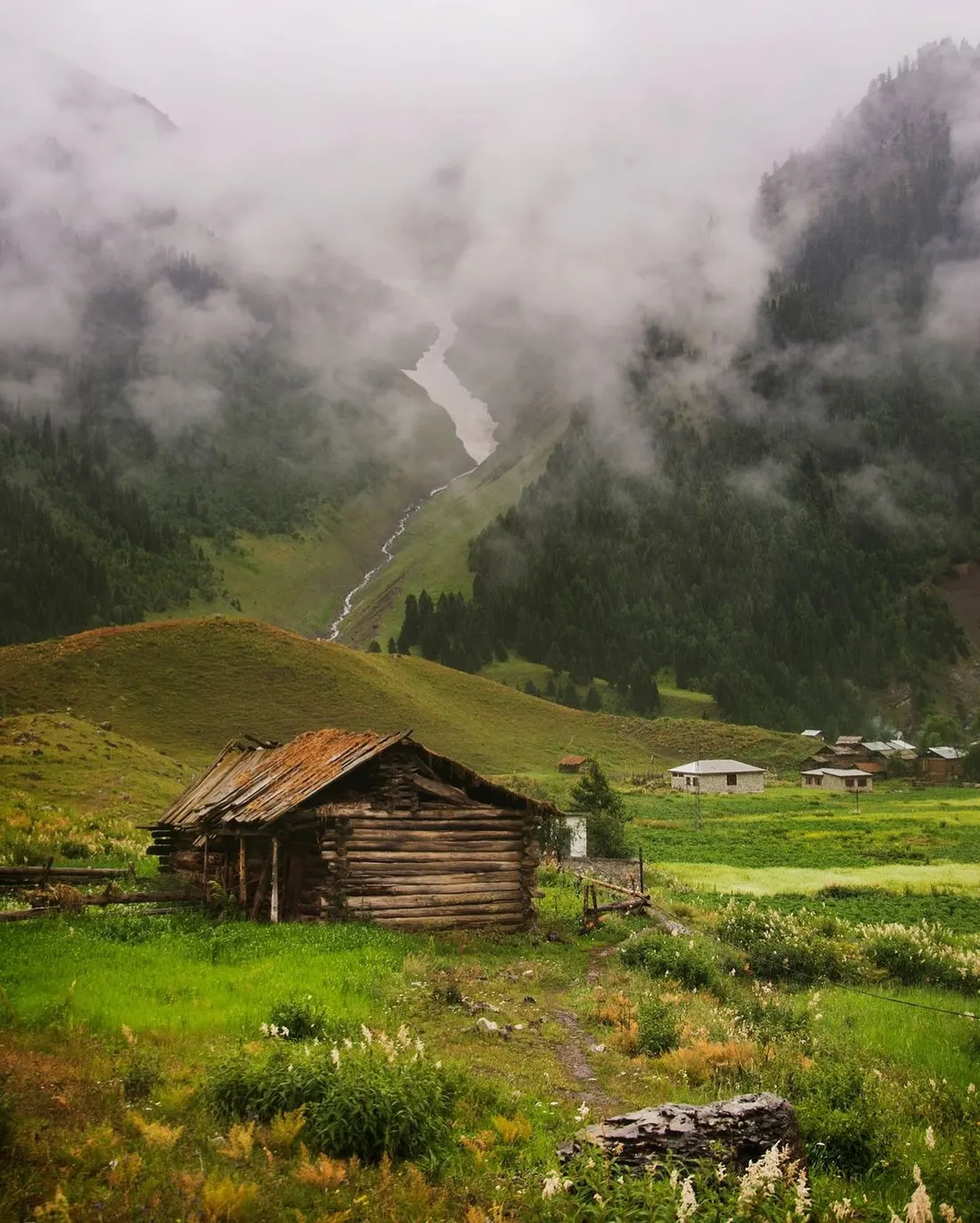 An old log cabin on the grass at Minimarg with village houses behind and cloud pouring off the forested ridges above