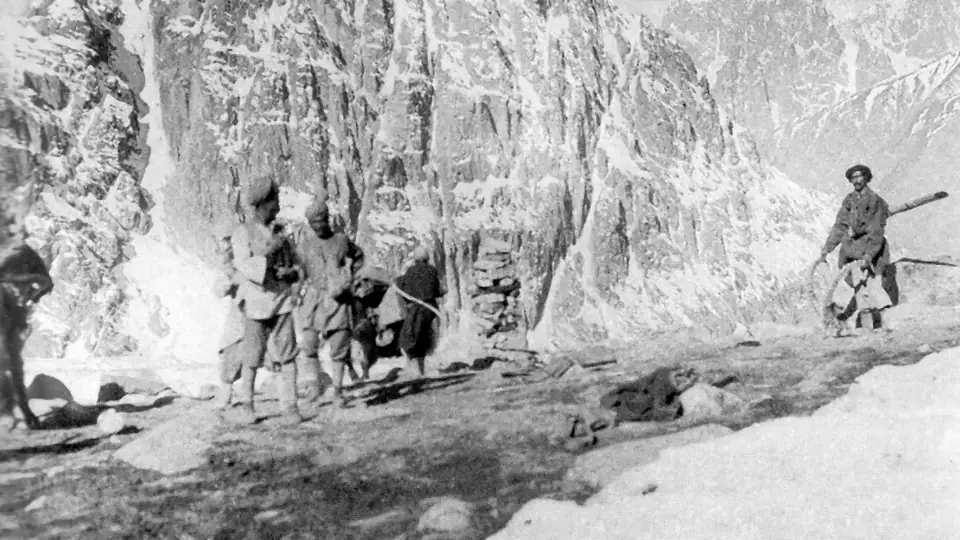 Fotografia in bianco e nero di viaggiatori e animali da soma sulla cima innevata del passo Mintaka nel 1918, con pareti di roccia e vette alle spalle, scattata da F. M. Bailey, Hunza, Pakistan