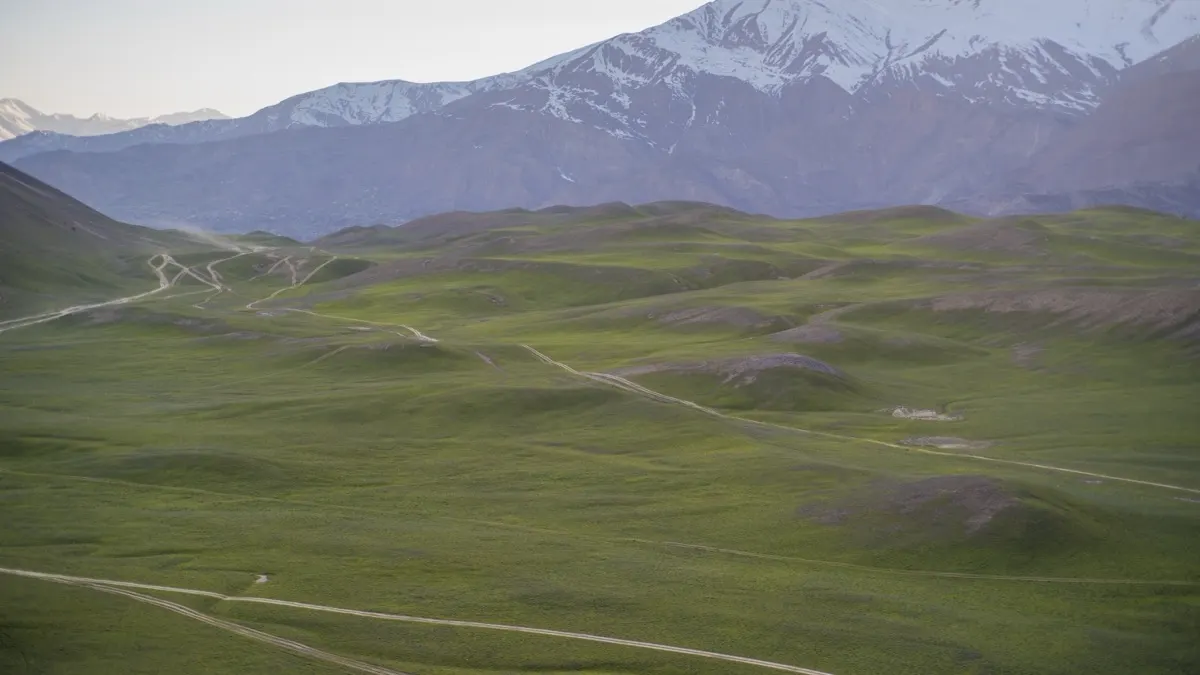 The empty green grass plateau of Qaqlasht Meadows in Upper Chitral with dirt tracks crossing it and the snow-covered Hindu Kush behind, Pakistan
