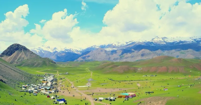 The Jashn-e-Qaqlasht festival encampment spread across the green Qaqlasht Meadows plateau in Upper Chitral, with the snow-covered Hindu Kush behind, Pakistan