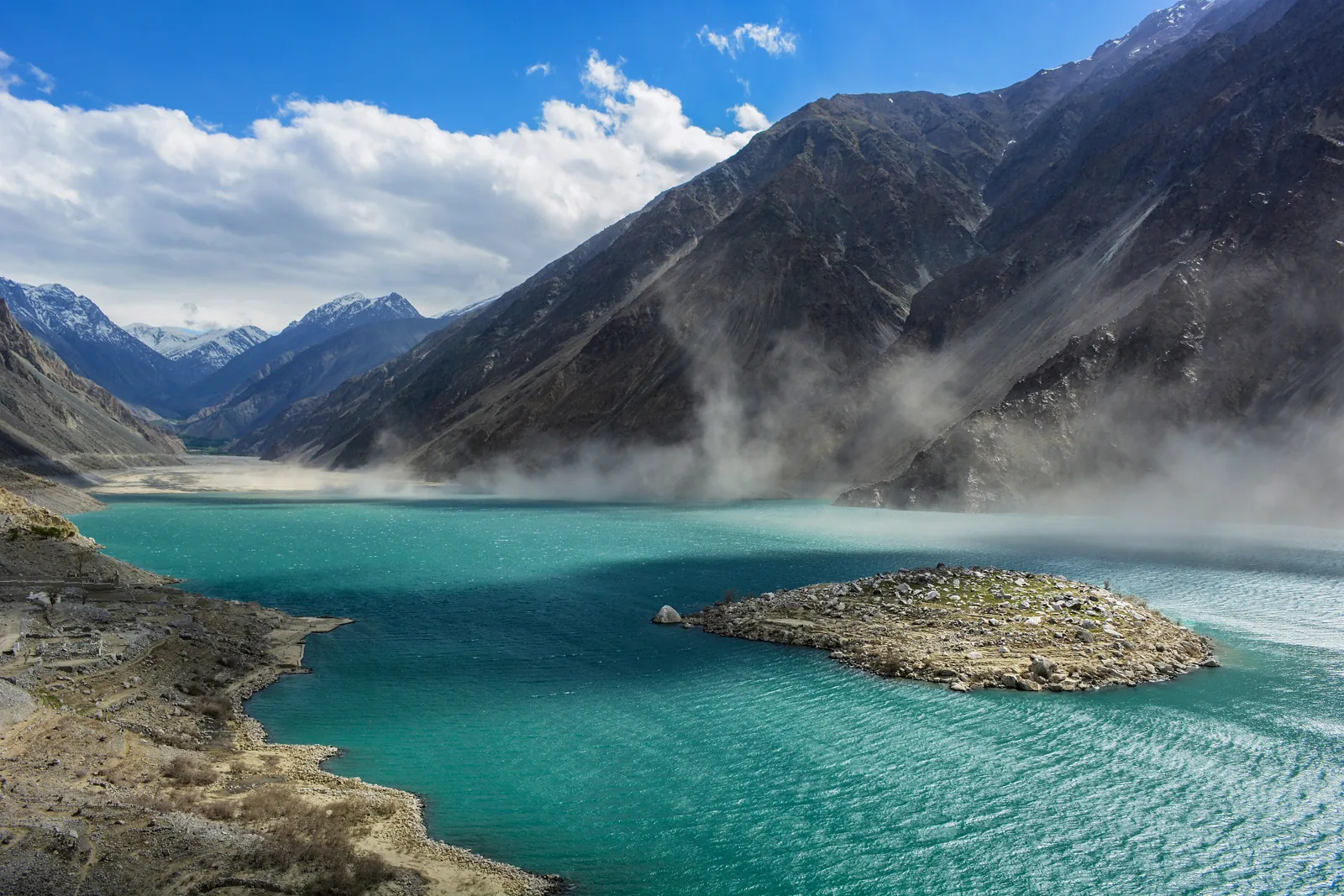 Turquoise Satpara Lake south of Skardu with its rocky island, as wind blows dust off the mountains