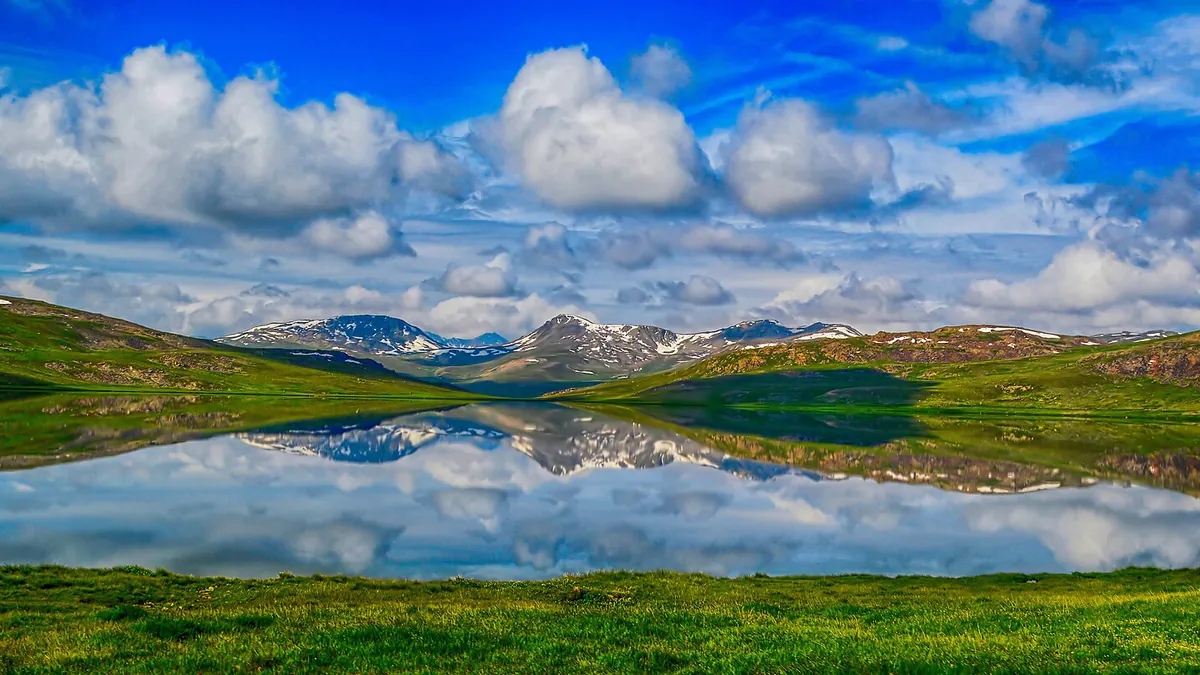 Sheosar Lake on the Deosai plateau reflecting white clouds and snowy hills, surrounded by green summer meadows