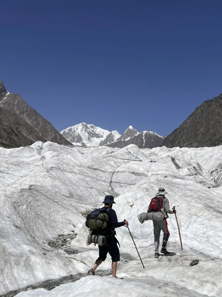 Zwei Trekker mit Rucksäcken und Trekkingstöcken auf dem blanken Eis des Passu-Gletschers, Karakorum-Gipfel voraus, Gojal, oberes Hunza