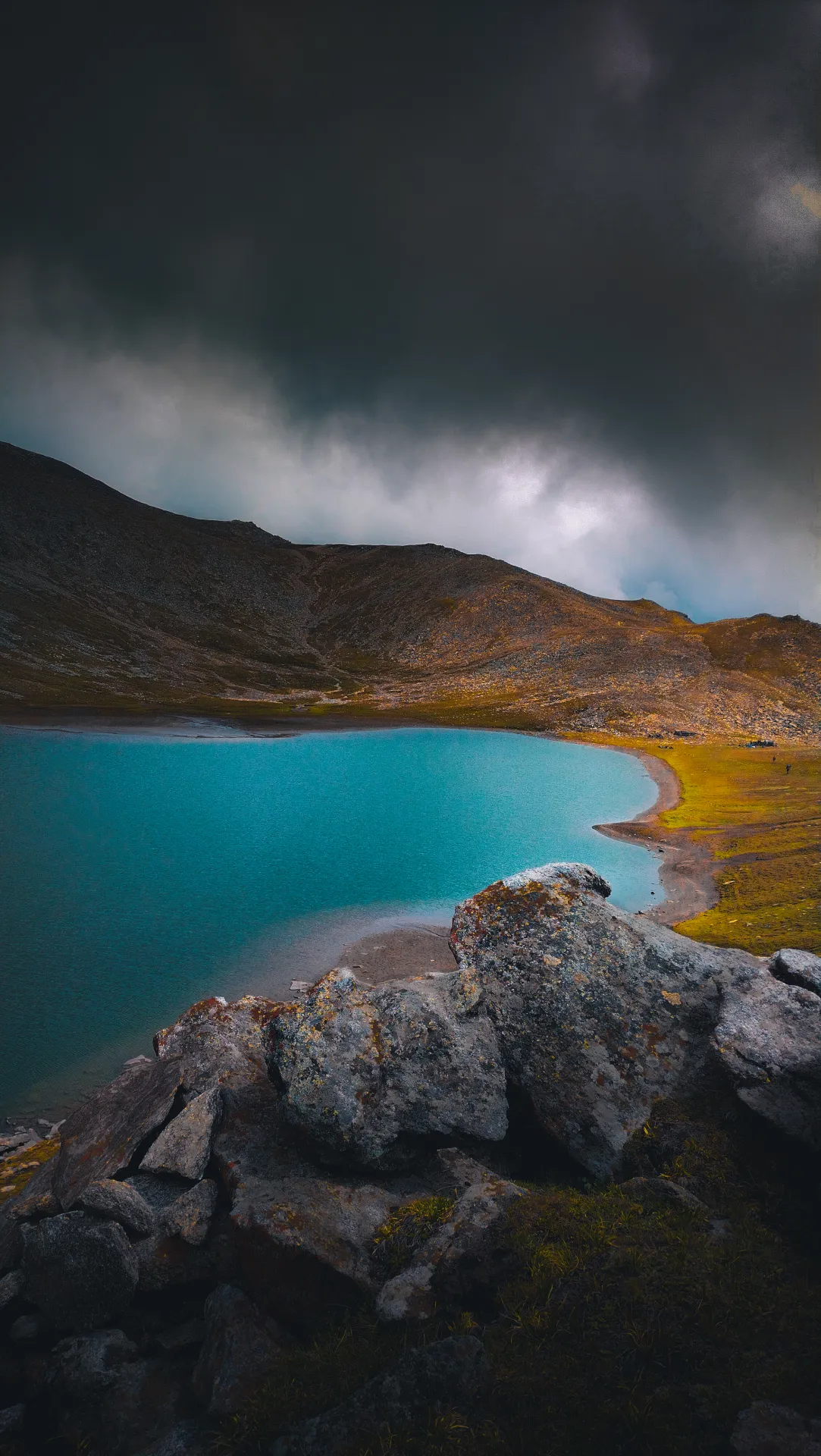Turquoise Rush Lake under dramatic storm clouds with granite boulders in the foreground, Nagar
